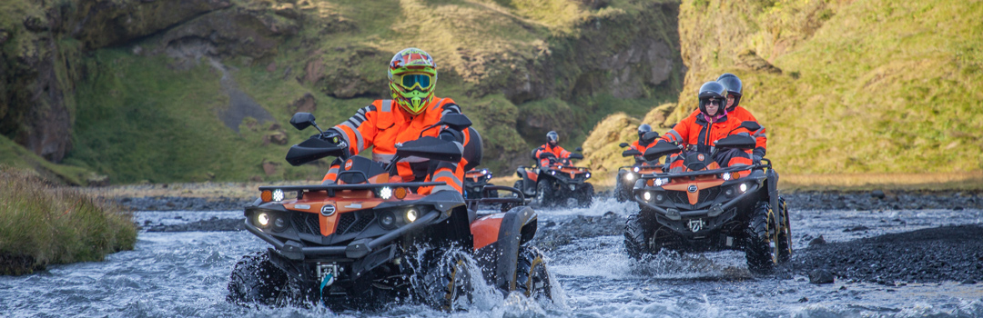 Group of people riding ATVs through a shallow river in a scenic Icelandic valley.